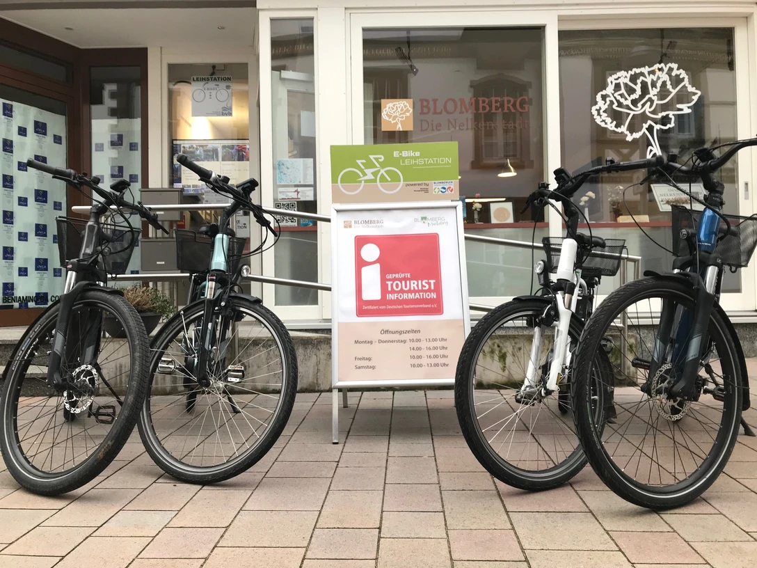 IMG_0570.jpg Vier E-Bikes stehen auf einer gepflasterten Fläche vor einem Tourist-Informationszentrum.