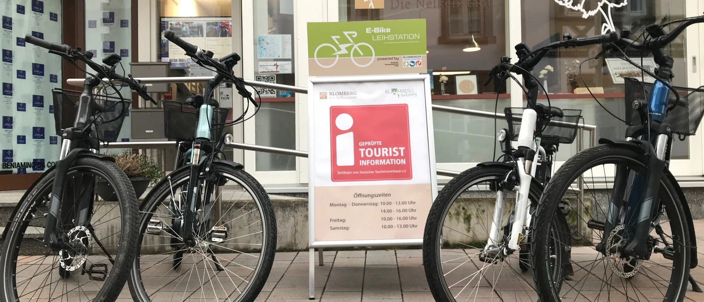 IMG_0570.jpg Vier E-Bikes stehen auf einer gepflasterten Fläche vor einem Tourist-Informationszentrum.