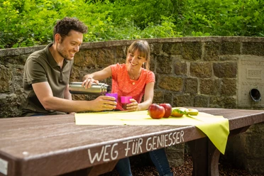 Werther-Arminiusquelle-Teutoburger-Wald-Tourismus-D-Ketz-077.jpg Ein Paar genießt ein Picknick an einem Holztisch im Grünen. Auf dem Tisch liegen Äpfel und Getränke.