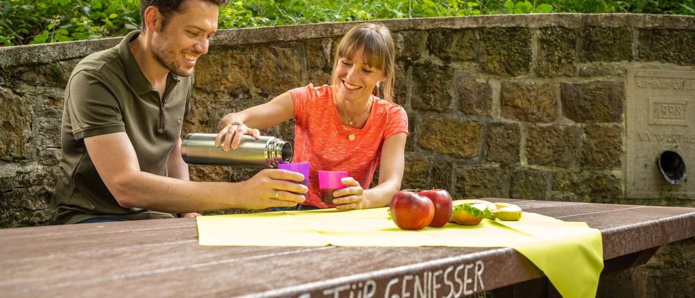 Ein Paar genießt ein Picknick an einem Holztisch im Grünen. Auf dem Tisch liegen Äpfel und Getränke.