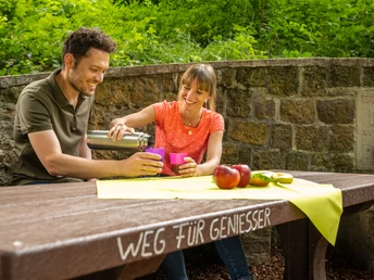 Werther-Arminiusquelle-Teutoburger-Wald-Tourismus-D-Ketz-077.jpg Ein Paar genießt ein Picknick an einem Holztisch im Grünen. Auf dem Tisch liegen Äpfel und Getränke.
