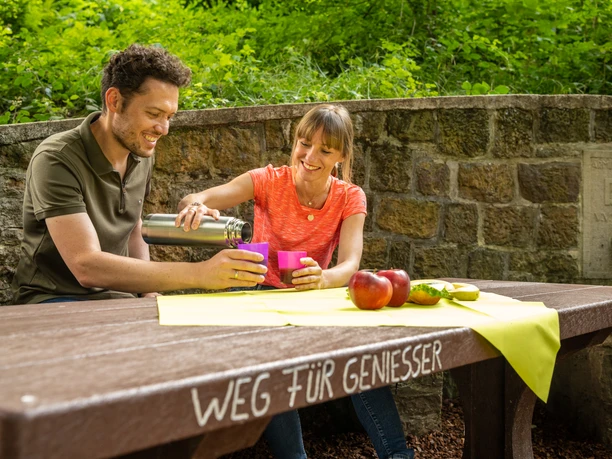 Werther-Arminiusquelle-Teutoburger-Wald-Tourismus-D-Ketz-077.jpg Ein Paar genießt ein Picknick an einem Holztisch im Grünen. Auf dem Tisch liegen Äpfel und Getränke.