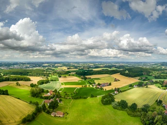 Weite, grüne Landschaften mit Feldern und Wäldern, verstreute Häuser unter einem bewölkten Himmel.