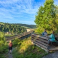 Zwei Personen sitzen an einem Tisch mit Blick auf den Altenbekener Viadukt im Grünen. Eine Wanderin läuft einen Pfad entlang.
