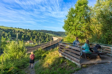 Altenbeken-Viadukt-Teutoburger-Wald-Tourismus-Patrick-Gawandtka-007.jpg Zwei Personen sitzen an einem Tisch mit Blick auf den Altenbekener Viadukt im Grünen. Eine Wanderin läuft einen Pfad entlang.