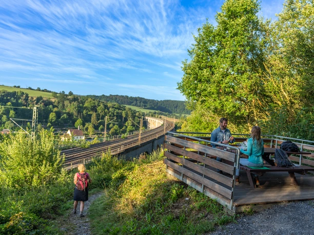 Altenbeken-Viadukt-Teutoburger-Wald-Tourismus-Patrick-Gawandtka-007.jpg Zwei Personen sitzen an einem Tisch mit Blick auf den Altenbekener Viadukt im Grünen. Eine Wanderin läuft einen Pfad entlang.