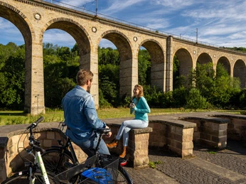 Ein Paar sitzt auf einer Bank vor dem beeindruckenden Altenbeken Viadukt unter blauem Himmel.