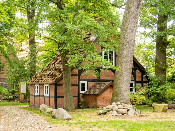 Fachwerkhaus mit roten Ziegeln, umgeben von hohen Bäumen und Natursteinen, an einem sonnigen Tag.