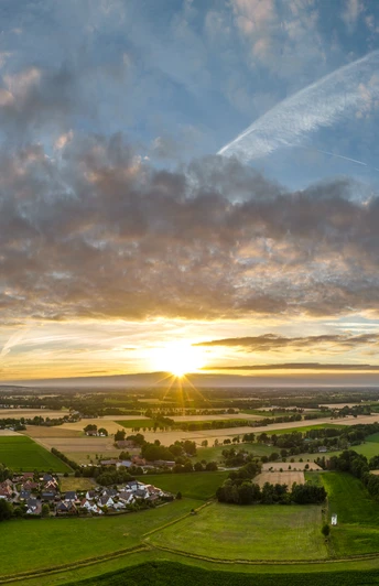 Sonnenuntergang über einer weiten, grünen Landschaft mit Feldern und Dörfern unter einem teils bewölkten Himmel.