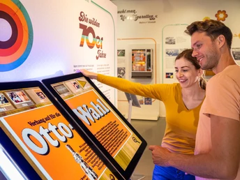 Ein Paar betrachtet interaktive Bildschirme in einer Ausstellung über die wilden 70er Jahre.
