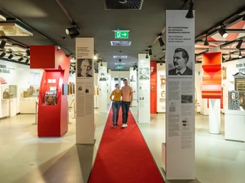 Ausstellungshalle mit roten Teppich, historischen Spielautomaten und Informationstafeln an weißen Säulen.