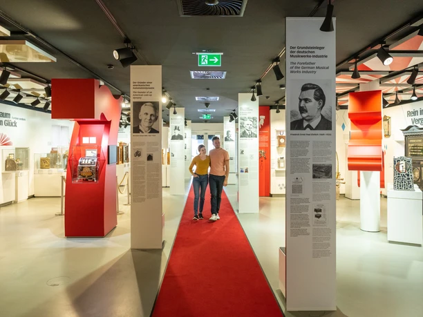 Espelkamp-Schloss Benkhausen-Automatenmuseum-Teutoburger-Wald-Tourismus-D-Ketz-077.jpg Ausstellungshalle mit roten Teppich, historischen Spielautomaten und Informationstafeln an weißen Säulen.