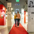 Ein Paar spaziert auf rotem Teppich durch ein modernes, lichtdurchflutetes Museum mit Infotafeln.