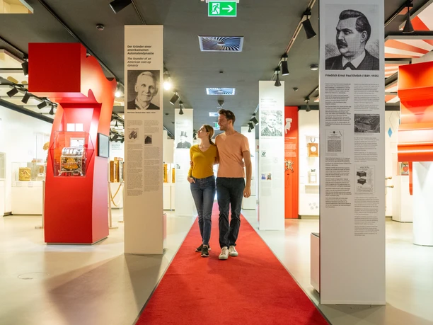 Ein Paar spaziert auf rotem Teppich durch ein modernes, lichtdurchflutetes Museum mit Infotafeln.