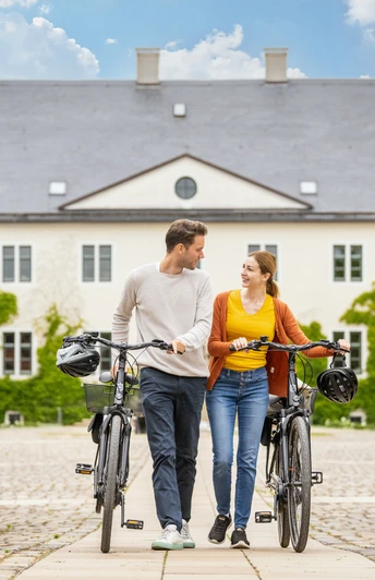 Ein Paar schiebt Fahrräder durch ein elegantes Tor vor einem historischen Gebäude bei Sonnenschein.