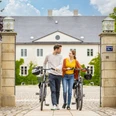 Espelkamp-Schloss Benkhausen-Teutoburger-Wald-Tourismus-D-Ketz-086.jpg Ein Paar schiebt Fahrräder durch ein elegantes Tor vor einem historischen Gebäude bei Sonnenschein.