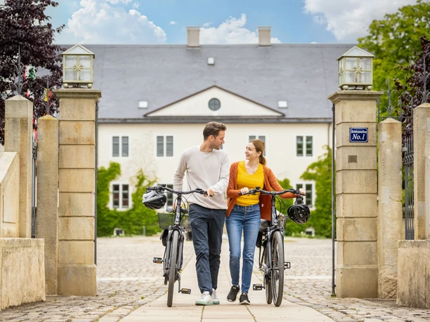 Espelkamp-Schloss Benkhausen-Teutoburger-Wald-Tourismus-D-Ketz-086.jpg Ein Paar schiebt Fahrräder durch ein elegantes Tor vor einem historischen Gebäude bei Sonnenschein.