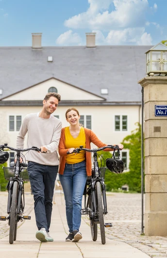 Ein junges Paar schiebt lächelnd Fahrräder durch ein steinernes Tor vor einem historischen Gebäude.