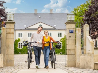 Espelkamp-Schloss Benkhausen-Teutoburger-Wald-Tourismus-D-Ketz-087.jpg Ein junges Paar schiebt lächelnd Fahrräder durch ein steinernes Tor vor einem historischen Gebäude.