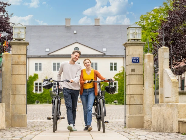Espelkamp-Schloss Benkhausen-Teutoburger-Wald-Tourismus-D-Ketz-087.jpg Ein junges Paar schiebt lächelnd Fahrräder durch ein steinernes Tor vor einem historischen Gebäude.