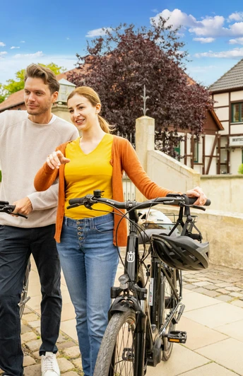 Espelkamp-Schloss Benkhausen-Teutoburger-Wald-Tourismus-D-Ketz-088.jpg Ein Paar mit Fahrrädern posiert lächelnd vor einem traditionellen Fachwerkgebäude in einem sonnigen Dorf.