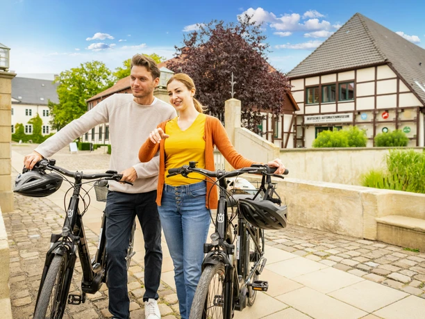 Espelkamp-Schloss Benkhausen-Teutoburger-Wald-Tourismus-D-Ketz-088.jpg Ein Paar mit Fahrrädern posiert lächelnd vor einem traditionellen Fachwerkgebäude in einem sonnigen Dorf.
