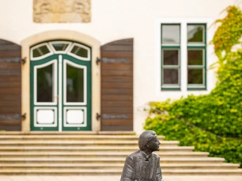 Bronzestatue eines sitzenden Mannes in Espelkamp vor einem Gebäude mit grünen Fensterrahmen.