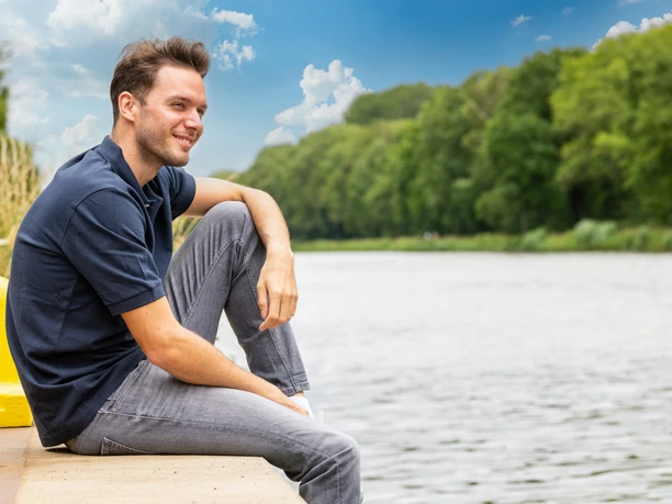 Ein Mann sitzt entspannt an einem Flussufer, umgeben von grünen Bäumen und blauem Himmel.
