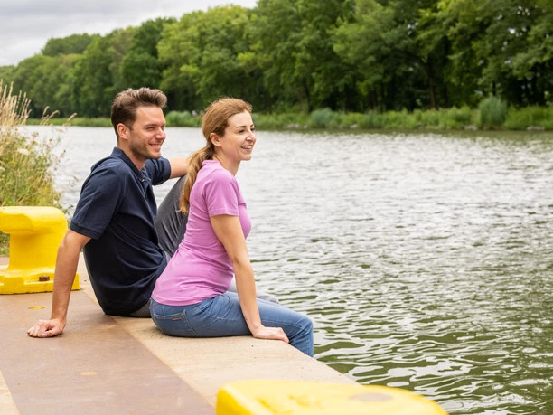 Ein Paar sitzt entspannt am Ufer eines Flusses, umgeben von grünen Bäumen, an einem bewölkten Tag.