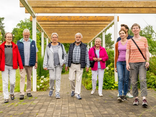 Espelkamp-Stadtführung-Teutoburger-Wald-Tourismus-D-Ketz-114.jpg Eine Gruppe älterer Menschen und ein Betreuer gehen gemeinsam unter einer Holzpergola im Park spazieren.