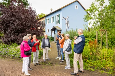Espelkamp-Stadtführung-Teutoburger-Wald-Tourismus-D-Ketz-115.jpg Eine Gruppe älterer Menschen auf einem Stadtrundgang in Espelkamp, vor einem blau gestrichenen Haus mit Kunstwerk an der Wand, umgeben von Pflanzen.