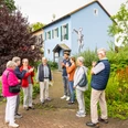 Espelkamp-Stadtführung-Teutoburger-Wald-Tourismus-D-Ketz-115.jpg Eine Gruppe älterer Menschen auf einem Stadtrundgang in Espelkamp, vor einem blau gestrichenen Haus mit Kunstwerk an der Wand, umgeben von Pflanzen.