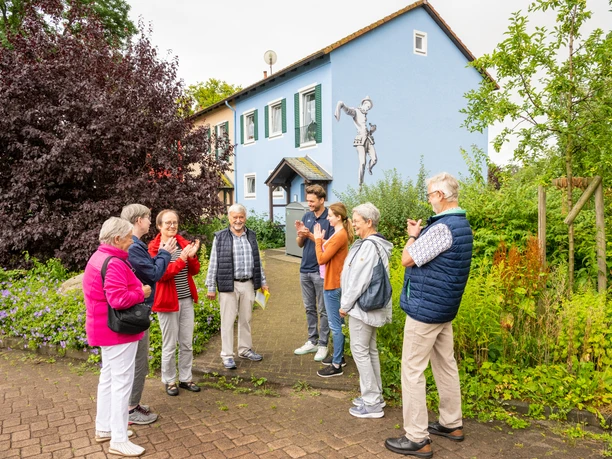 Espelkamp-Stadtführung-Teutoburger-Wald-Tourismus-D-Ketz-115.jpg Eine Gruppe älterer Menschen auf einem Stadtrundgang in Espelkamp, vor einem blau gestrichenen Haus mit Kunstwerk an der Wand, umgeben von Pflanzen.