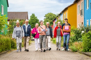 Eine Gruppe fröhlicher Menschen spaziert gelassen auf einem Weg, umgeben von bunten Häusern.