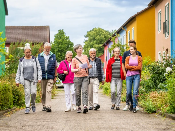Espelkamp-Stadtführung-Teutoburger-Wald-Tourismus-D-Ketz-118.jpg Eine Gruppe fröhlicher Menschen spaziert gelassen auf einem Weg, umgeben von bunten Häusern.