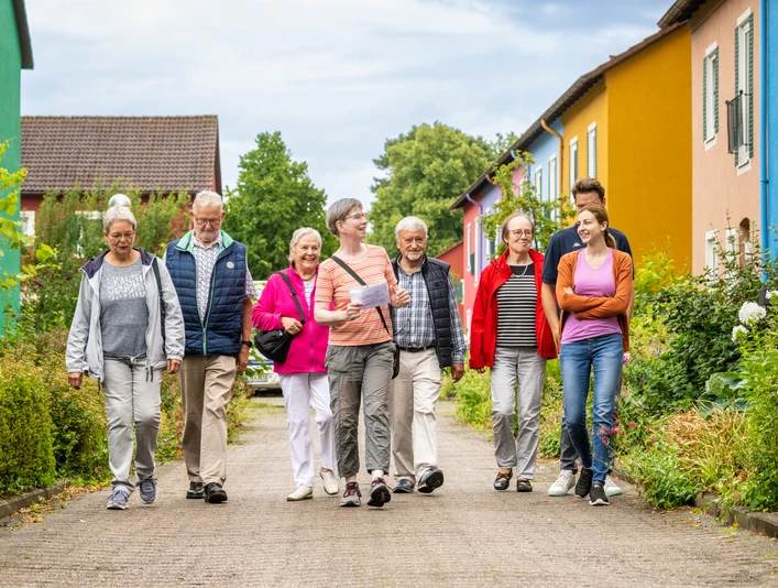 Espelkamp-Stadtführung-Teutoburger-Wald-Tourismus-D-Ketz-118.jpg Eine Gruppe fröhlicher Menschen spaziert gelassen auf einem Weg, umgeben von bunten Häusern.