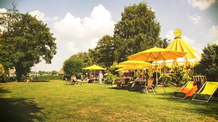 Schirmbar (Jens Butz).jpg Blick auf eine Wiese mit mehreren Sitzplätzen, gelben Sonnenschirmen und Menschen, die dort zusammensitzen und sich unterhalten