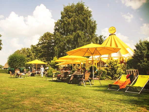 Schirmbar (Jens Butz).jpg Blick auf eine Wiese mit mehreren Sitzplätzen, gelben Sonnenschirmen und Menschen, die dort zusammensitzen und sich unterhalten
