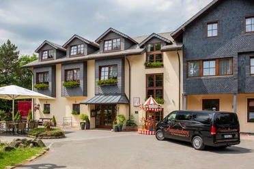 Augustusberg - Hotel & Restaurant Three-storey building with wooden shingle roofs, next to it a decorative circus wagon.