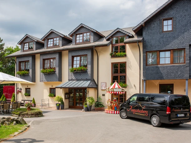 Augustusberg - Hotel & Restaurant Dreistöckiges Gebäude mit hölzernen Schindeldächern, daneben ein dekorativer Zirkuswagen.