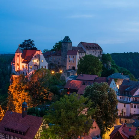 Burg Hohnstein am Abend Burg auf einem Hügel bei Dämmerung, umgeben von beleuchteten Fachwerkhäusern und Bäumen, mit einem bewaldeten Hintergrund.