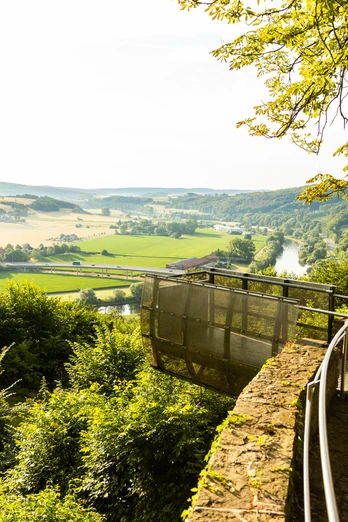 Aussichtsplattform mit Blick auf eine idyllische Landschaft aus Feldern, Hügeln und einem Fluss.