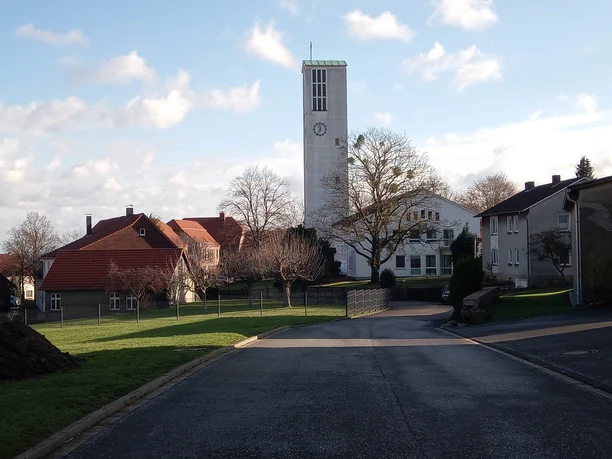 Nettelstedt Kirchturm mit Kreuz, umgeben von Wohnhäusern in Nettelstedt, bei sonnigem Himmel und wenigen Wolken.