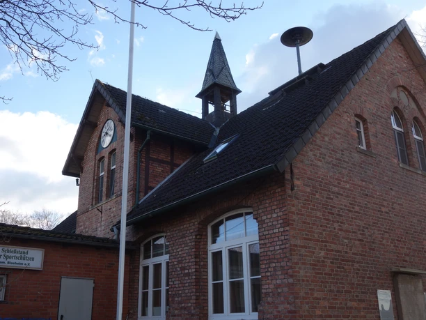 Obermehnen Schule Backsteingebäude mit Uhrturm und spitzem Dach, umgeben von kahlen Bäumen unter blauem Himmel.