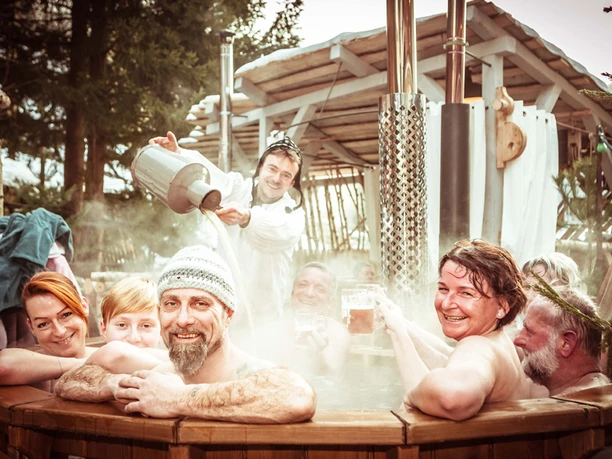 Group of people relaxing in a steaming wooden tub outdoors while one person pours hot water; cheerful mood.