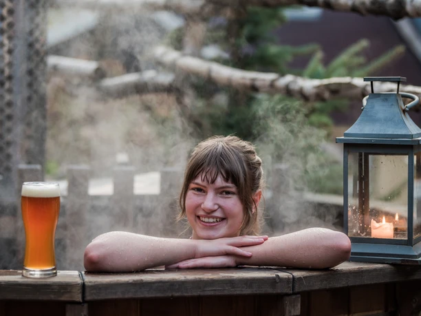 A woman smiles in a steaming wooden tub, next to a glass of beer and a burning candle in a lantern.