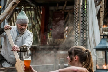 A man with a gray beard and cap stirs with a wooden stick in a steaming vat while a woman holds a glass of beer.