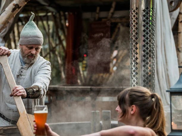 Badezuberei Schmilka Ein Mann mit grauem Bart und Mütze rührt mit einem Holzstab in einem dampfenden Bottich, während eine Frau ein Bierglas hält.