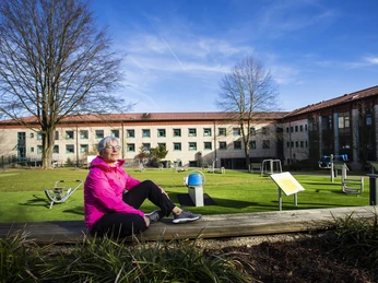 Outdoor Workpark Klinik Berlin Ältere Frau entspannt auf Holzbalken vor Klinik, umgeben von Fitnessgeräten und blauem Himmel.