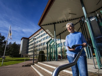Ältere Frau mit Broschüre vor Klinikgebäude in Bad Driburg, sonniger Tag, blauer Himmel.